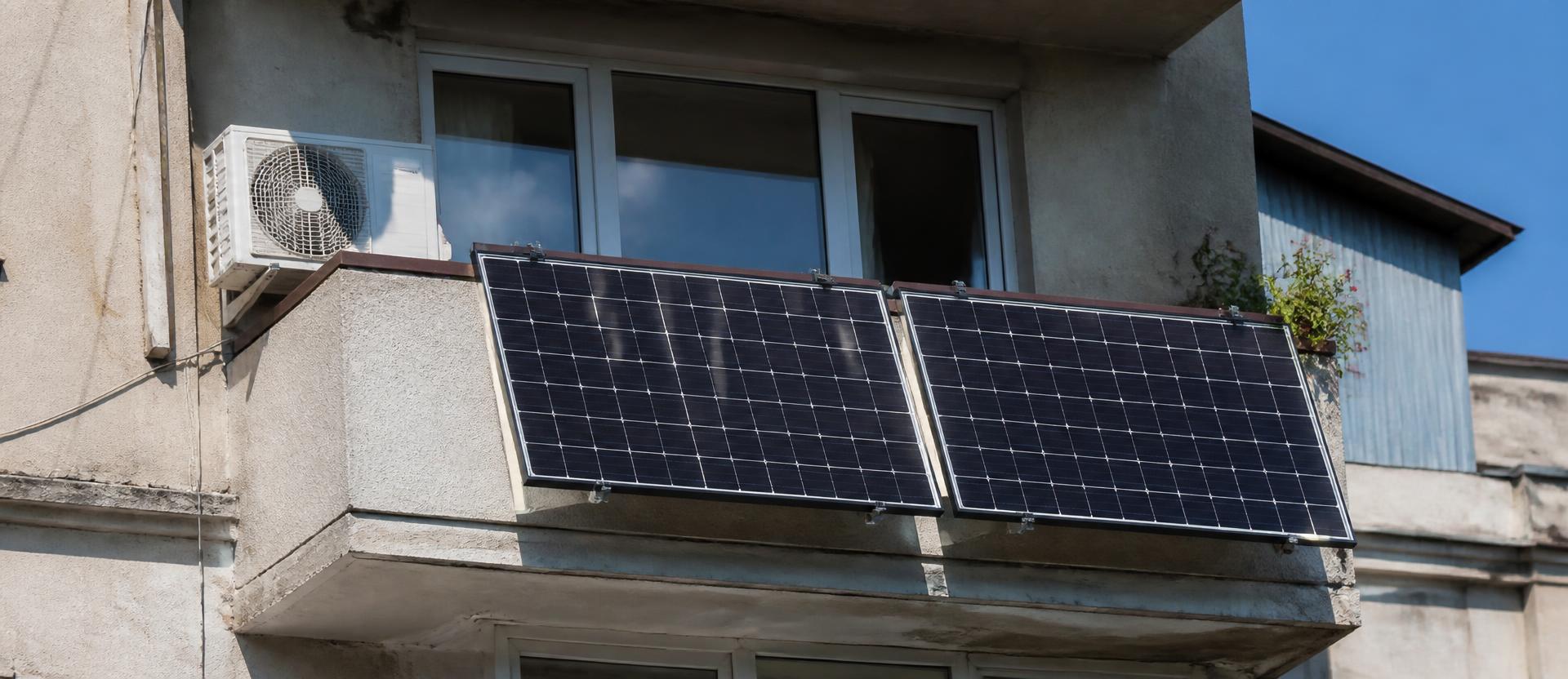 Unitate exterioară de aer condiționat montată lângă un balcon de bloc din România, cu două panouri solare fixate pe balustradă, în plin soare de vară