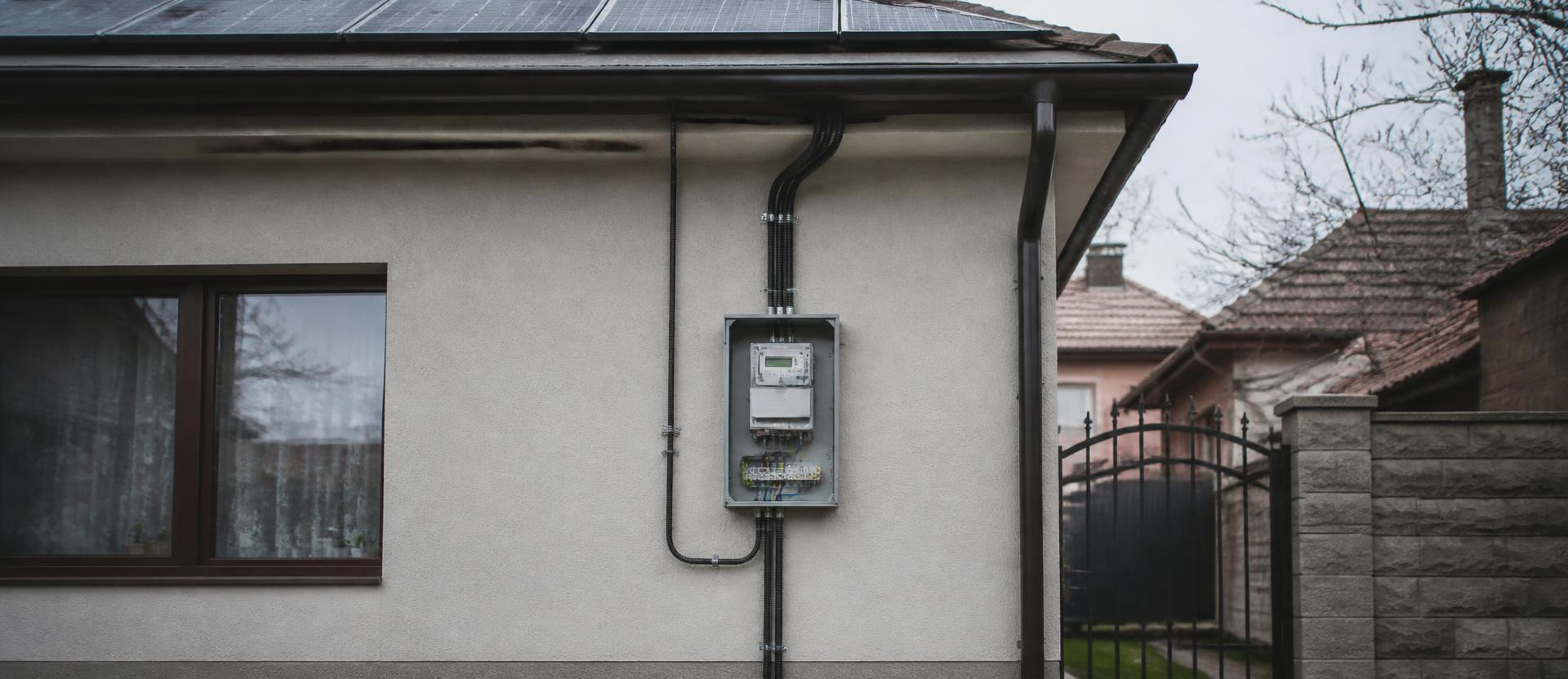 Cutie de contor electric pe fațada unei case din România, cu panouri solare pe acoperiș și racord către rețea