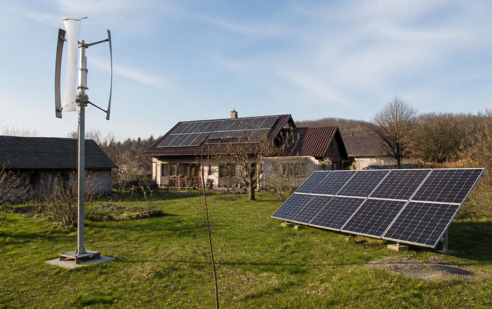 Comparație vizuală într-o curte de casă din România: turbină eoliană verticală mică pe stâlp și panouri solare montate la sol, cu casa având panouri și pe acoperiș