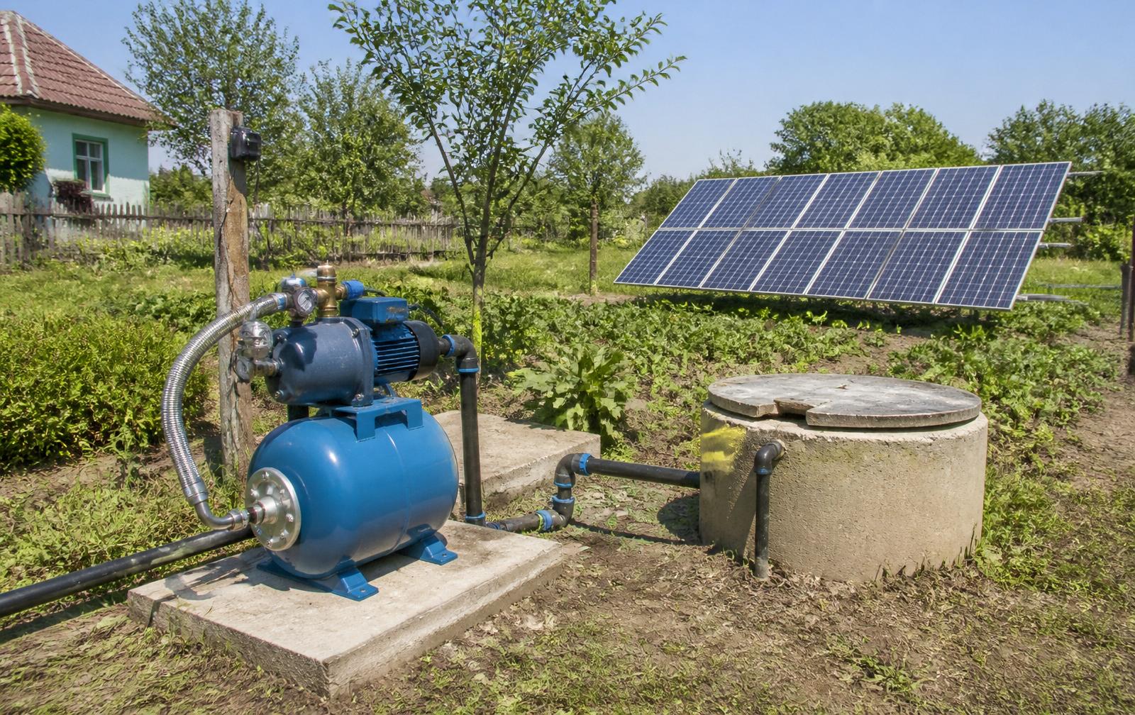 Grup de hidrofor cu rezervor de presiune și pompă instalate pe soclu de beton lângă gura unui puț, cu panouri fotovoltaice montate la sol în grădină